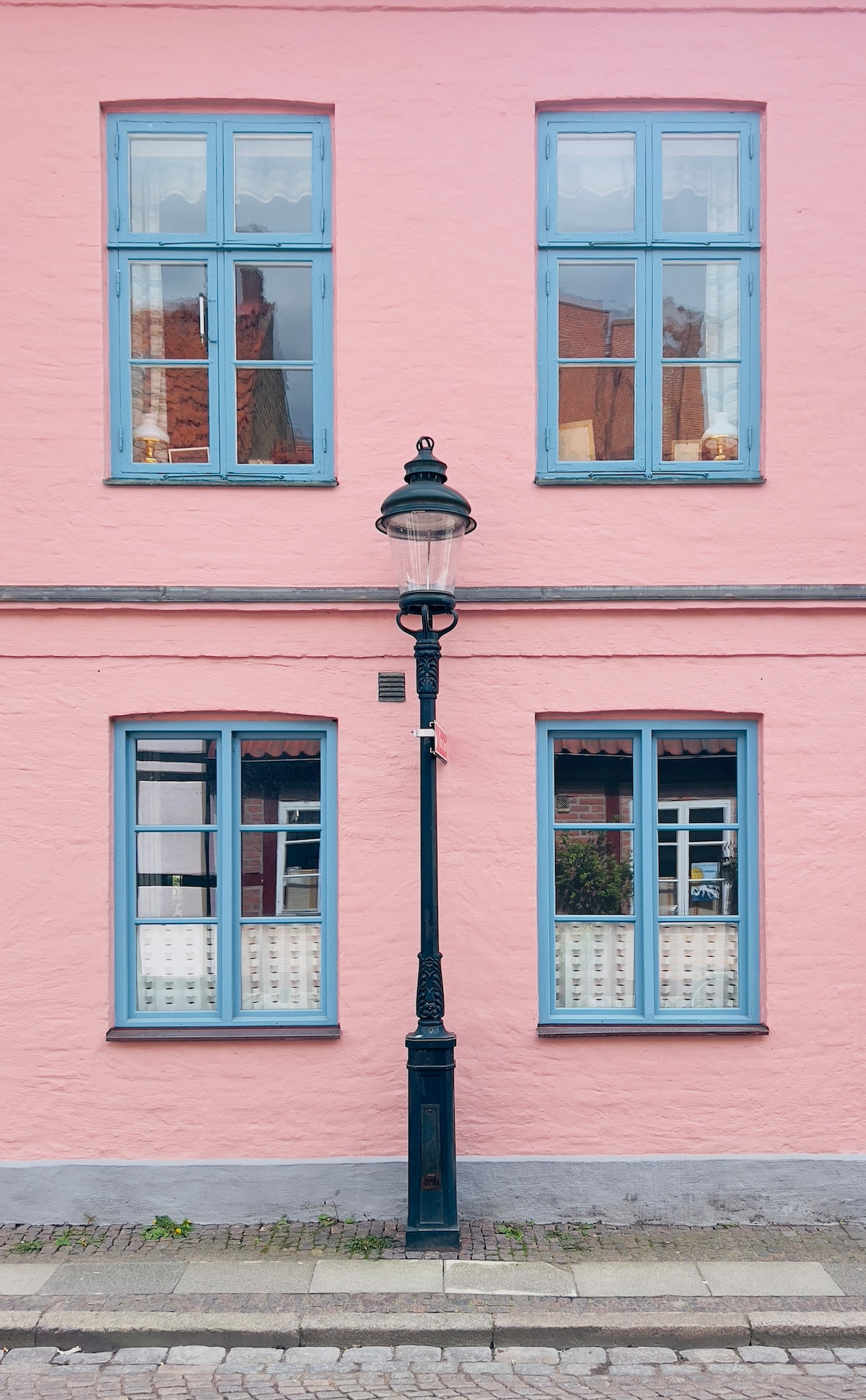 A charming pink building features four windows with blue frames, reflecting the character of the area. A lantern pole stands centrally in front, enhancing the quaint street view. The cobblestone path complements the architectural style, contributing to the inviting atmosphere of Lund.