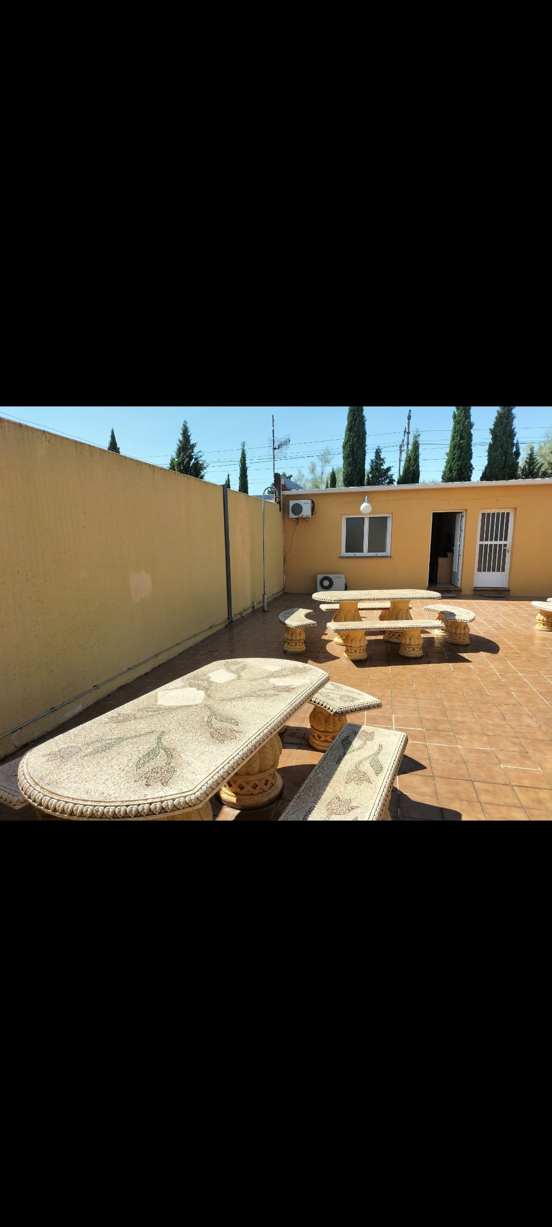 An outdoor terrace features several stone tables and benches arranged neatly along the tiled floor. The surrounding walls are painted in a soft yellow tone, with greenery visible through the tall trees beyond. An air conditioning unit is mounted on the wall of the adjoining building.
