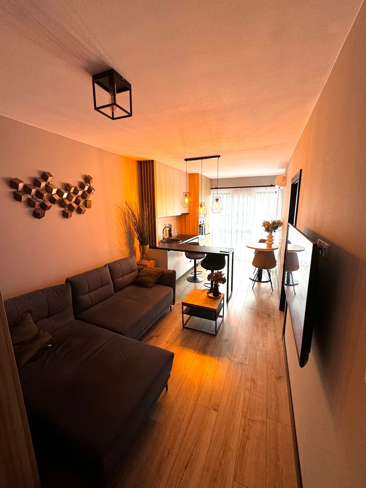 A modern living area is seen with a sectional sofa arranged against a wall. A small coffee table sits in front, while two stylish barstools are positioned at a counter in the background. Warm lighting enhances the contemporary design, with large windows allowing natural light.