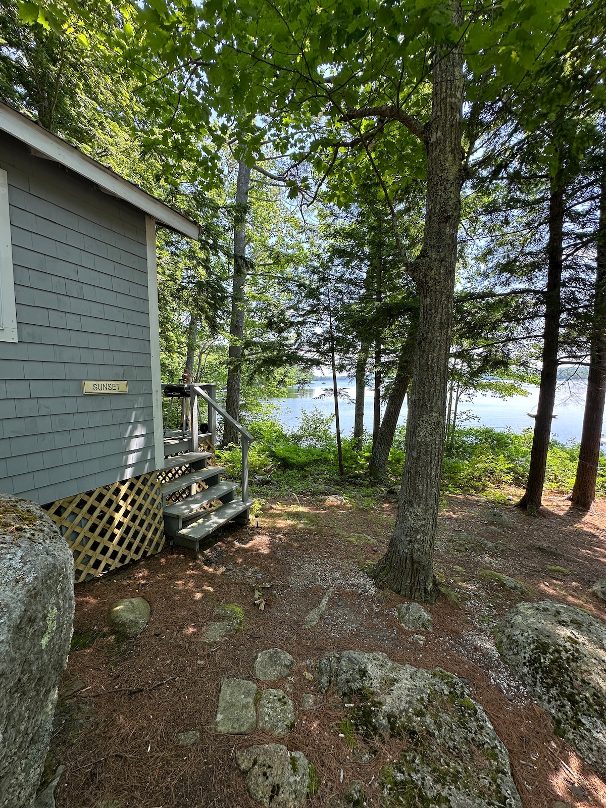 The exterior of a cozy cabin is framed by trees, showcasing a small set of steps leading to the entrance. Beyond the cabin, the serene lake is visible through the foliage, creating a peaceful natural setting.