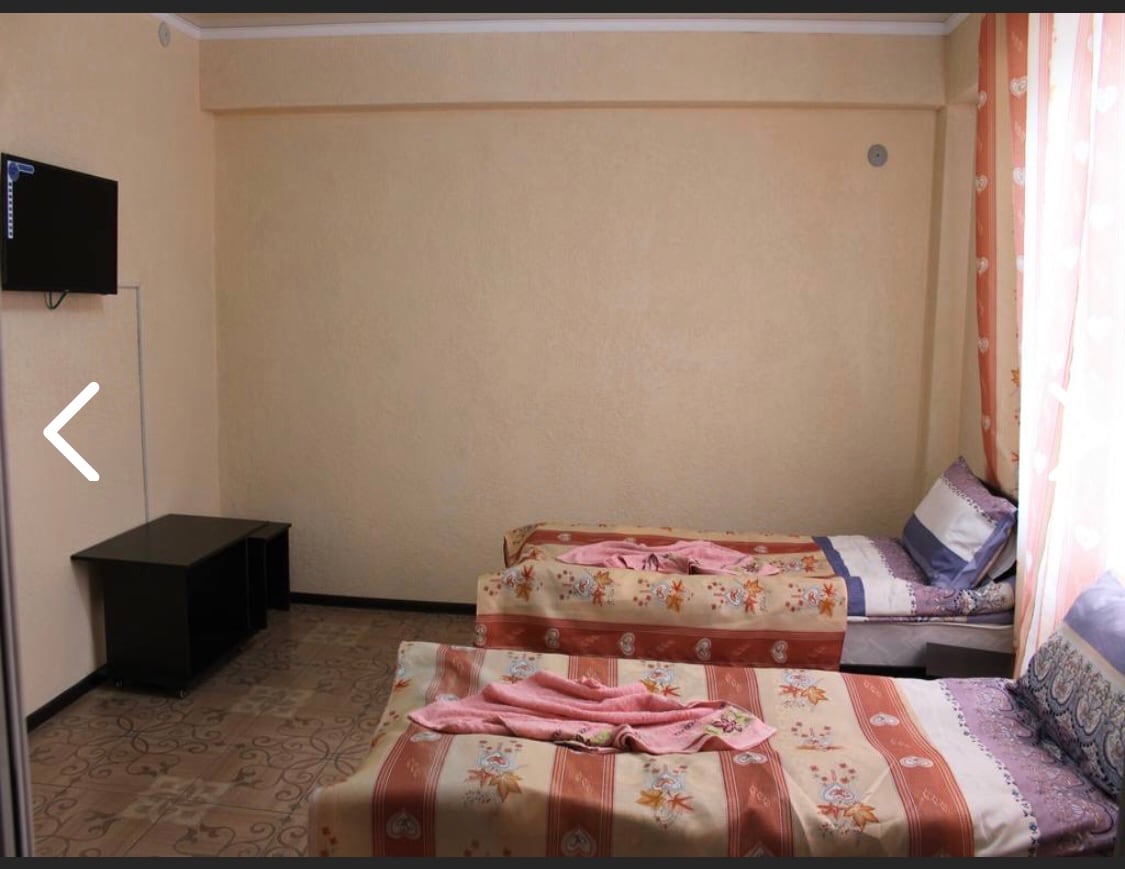 A room is furnished with two single beds, each dressed in floral patterned covers. A small dark wooden table sits against one wall, and a wall-mounted television is positioned opposite the beds. Soft sunlight filters through light curtains, creating a bright atmosphere.