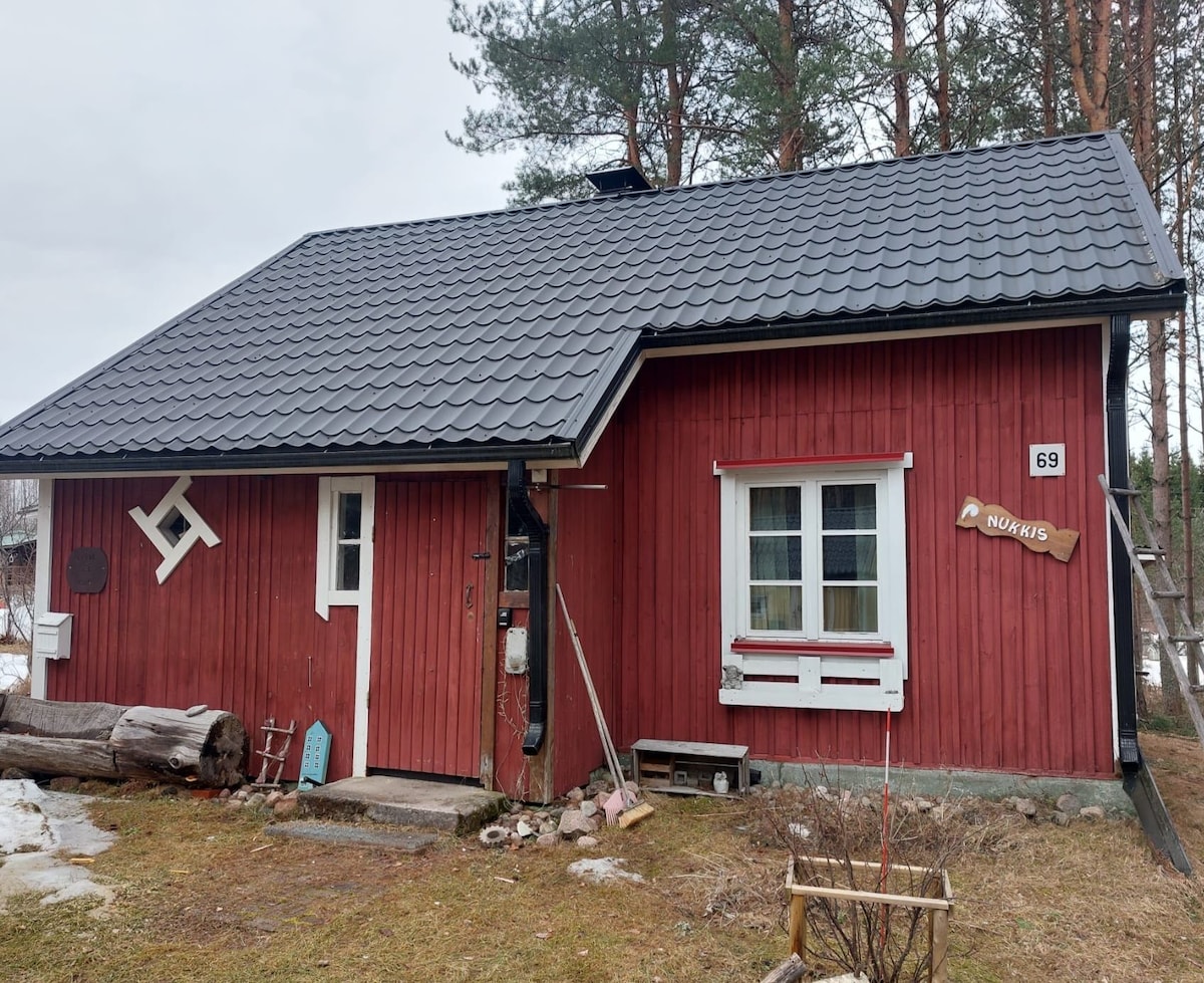 A red wooden cabin is framed by a grassy yard, with a sloped roof and white-trimmed windows. A wooden sign displaying the number '69' is attached to the wall, and logs are arranged casually in the foreground. Tall pine trees rise in the background.