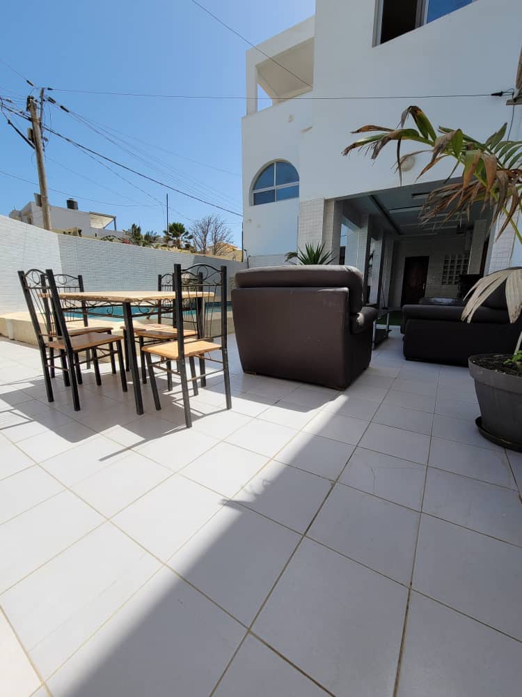 An outdoor seating area is presented with a dining table surrounded by four chairs. Brown leather sofas are positioned nearby, and the space is adorned with potted plants. Sunlight illuminates the tiled floor, creating a bright and open environment.