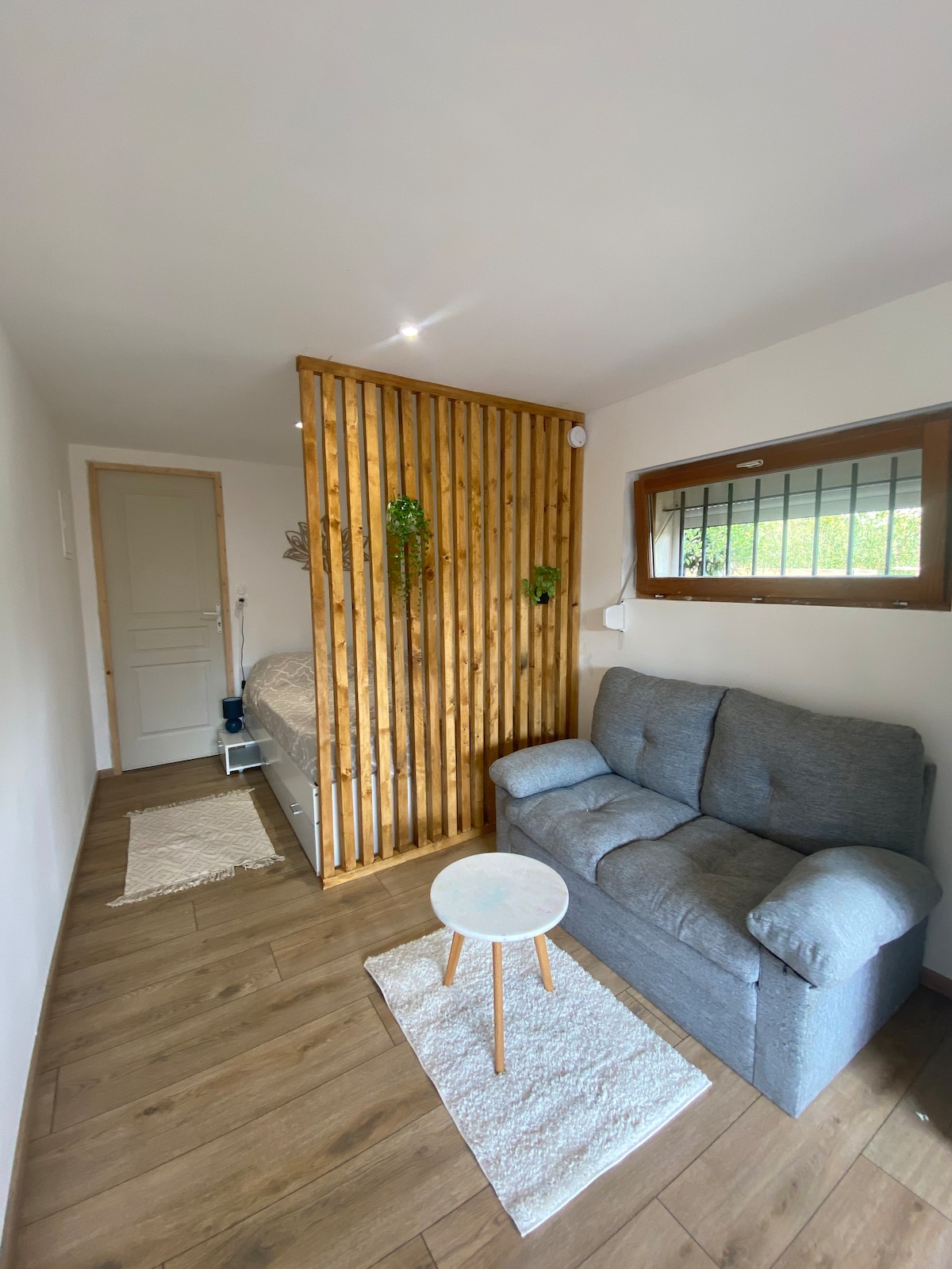 A cozy seating area features a gray couch positioned beside a small round table. Natural light enters through a window, illuminating the wood-paneled divider that separates the sleeping space. A neutral-colored rug lies on the wooden floor, adding warmth to the ambiance.