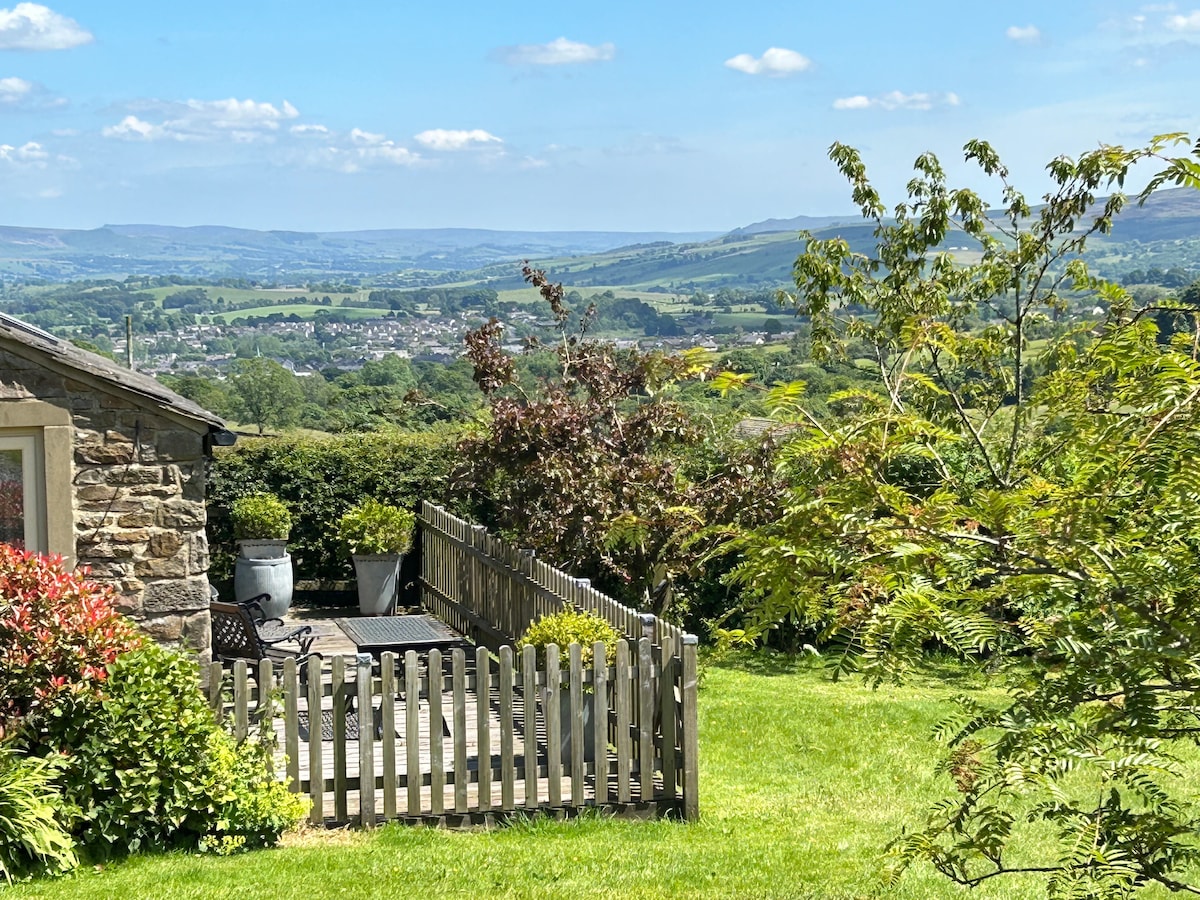 The image showcases a cottage patio area surrounded by greenery. A wooden fence encloses the space, which features outdoor seating and a table. The expansive countryside view includes rolling hills and a clear blue sky, creating a calming outdoor environment.