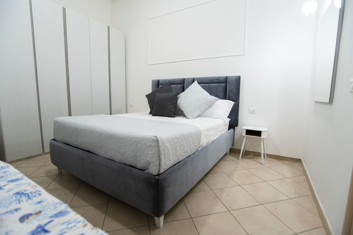 A spacious bedroom features a grey upholstered bed with multiple decorative pillows. A small nightstand is positioned beside the bed, and a large wardrobe occupies one wall. Soft, neutral tones create a calming ambiance.