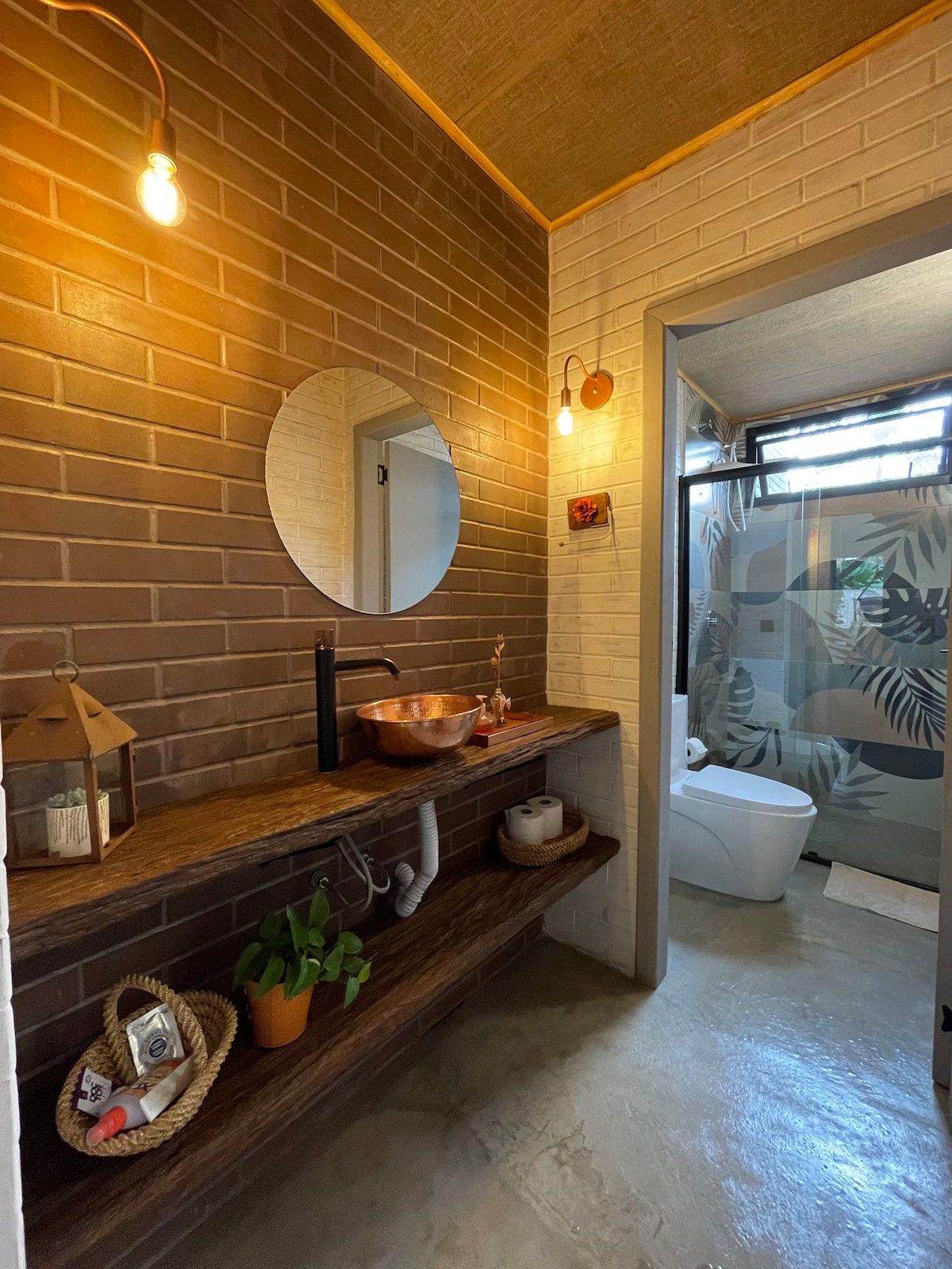 The bathroom features a spacious layout with warm brown brick walls and a round mirror above a wooden vanity. A copper sink is highlighted alongside a small plant. The area is well-lit with modern fixtures, leading to a separate shower space decorated with tropical patterns.