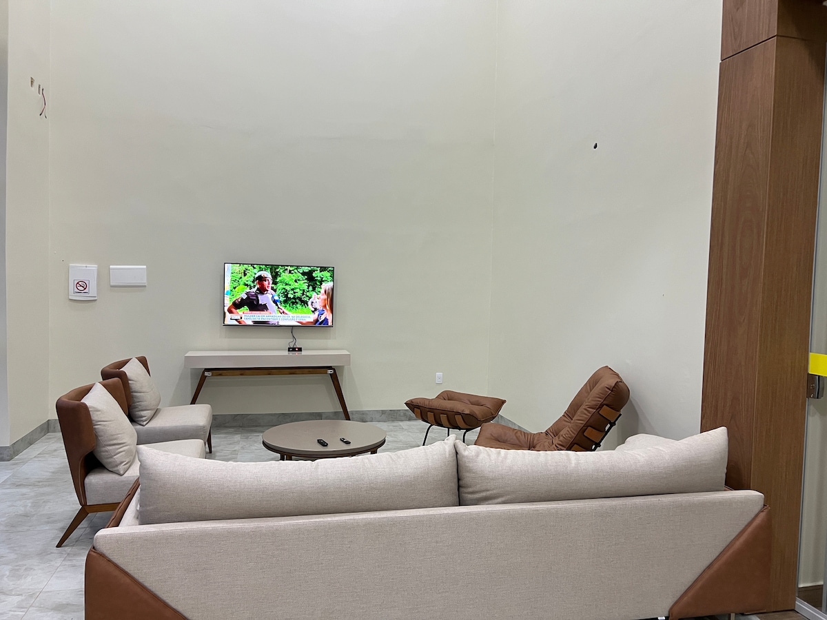 A cozy seating area is arranged with a light-colored sofa, two brown lounge chairs, and a circular coffee table. A television is mounted on the wall, displaying an outdoor scene. Soft gray flooring complements the neutral tones of the furniture and walls.