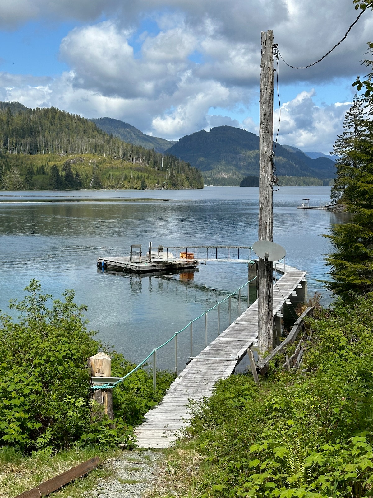 A wooden dock extends into calm waters, leading to a small floating platform. Lush greenery frames the scene, with mountains rising in the background under a partly cloudy sky. The tranquil setting suggests a secluded access point for boats.