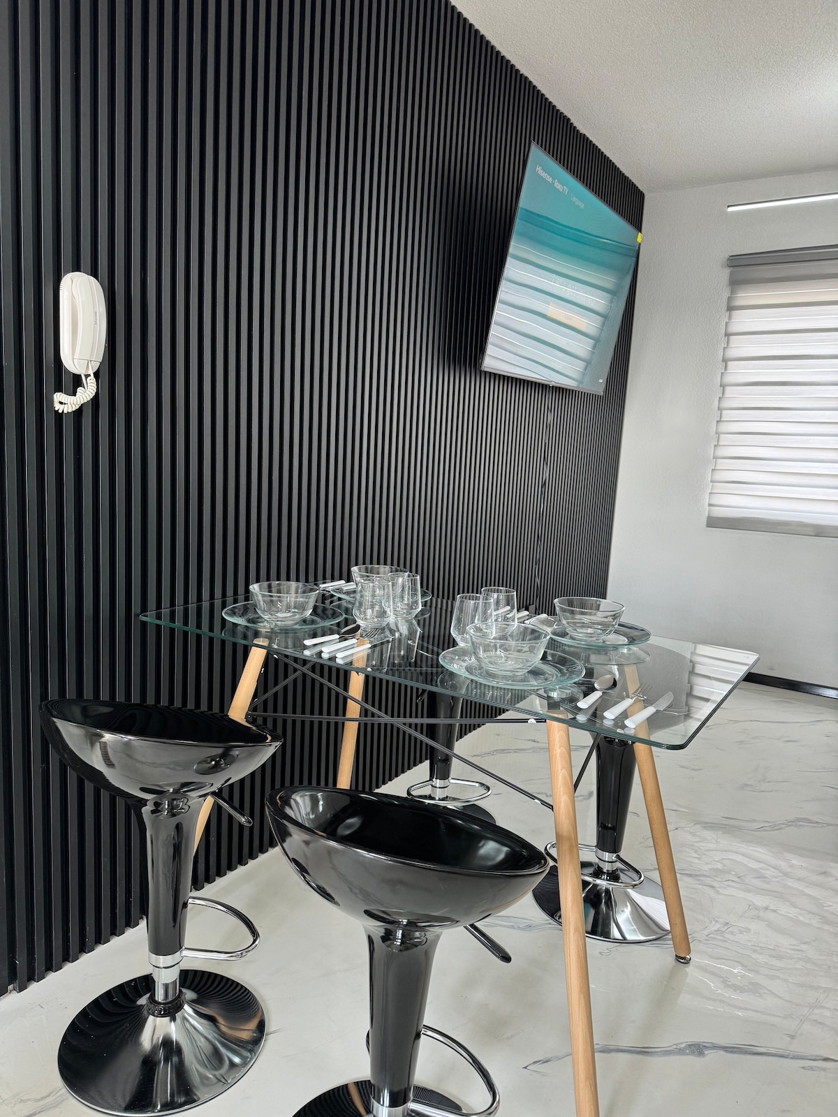 A modern dining area is showcased with a sleek glass table supported by wooden legs. Three high, glossy black stools surround the table. The background features textured black paneling, and a mounted television is visible above the table, providing a contemporary touch.