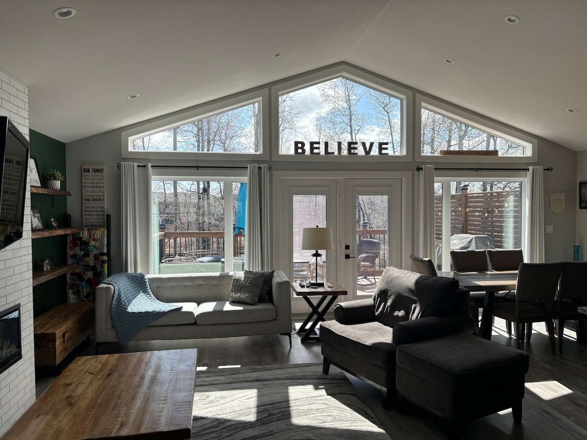 The open living space features large windows that fill the room with natural light. A comfortable sofa and armchairs are arranged around a wooden coffee table. Sliding doors lead to a deck with outdoor seating. Decorative elements include framed quotes on the walls.