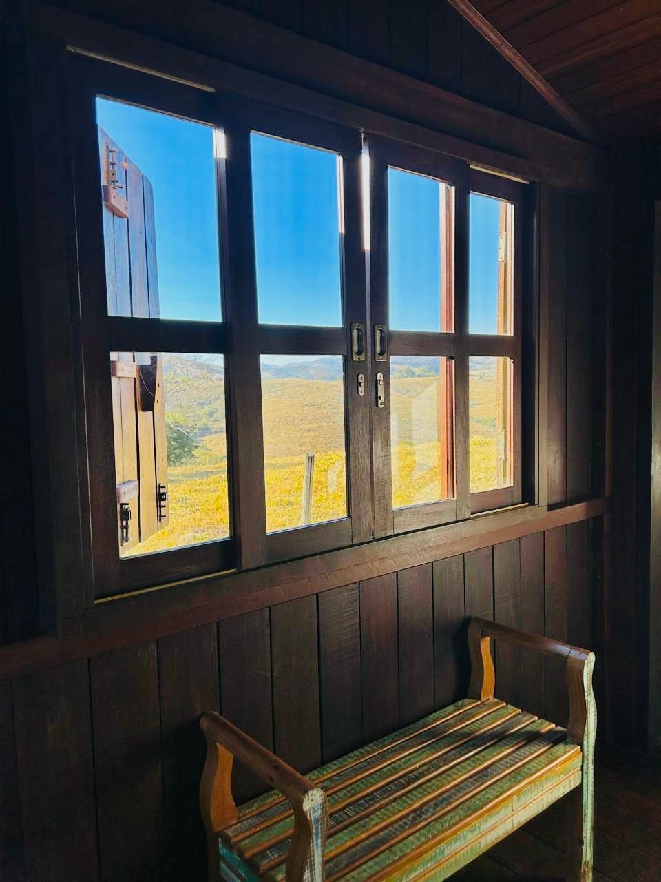 A wooden interior features a small striped bench beside a window, which frames a view of rolling hills bathed in natural light. The dark wood walls complement the scenic landscape outside, adding a sense of connection to nature.