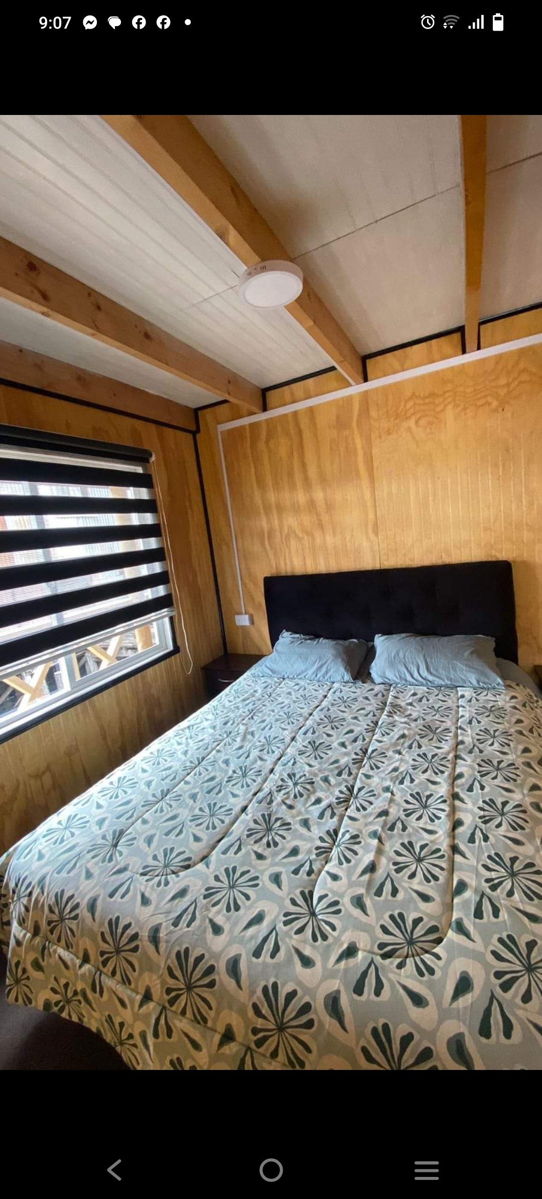 A cozy sleeping area features a dark upholstered headboard against wood-paneled walls. A bed with a patterned cover and two soft pillows is positioned near a window adorned with dual-layered shades, allowing natural light to filter through.