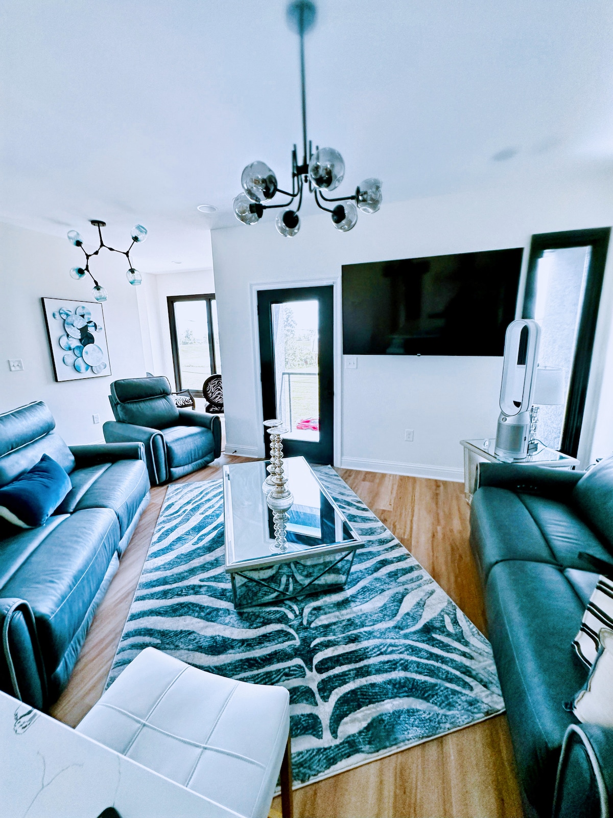 A cozy living area features sleek leather couches arranged around a glass coffee table on a patterned area rug. Large windows allow natural light, while a modern ceiling light and a mounted flat-screen TV enhance the contemporary ambiance.