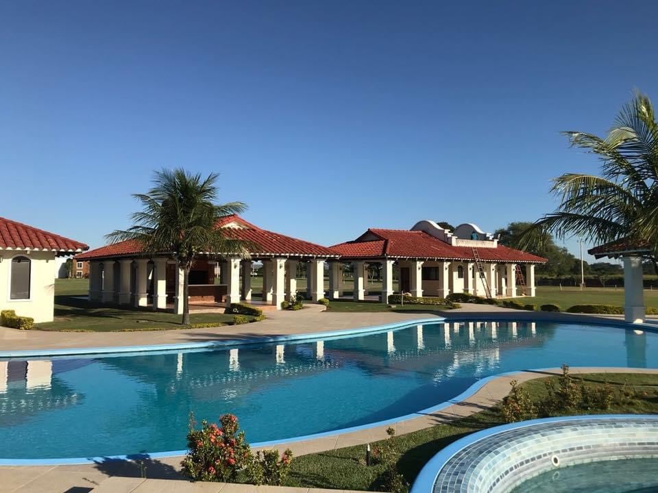 A serene outdoor area is highlighted by a large swimming pool, surrounded by well-maintained gardens and palm trees. Several inviting structures with red-tiled roofs can be seen in the background, offering shaded spaces for relaxation.