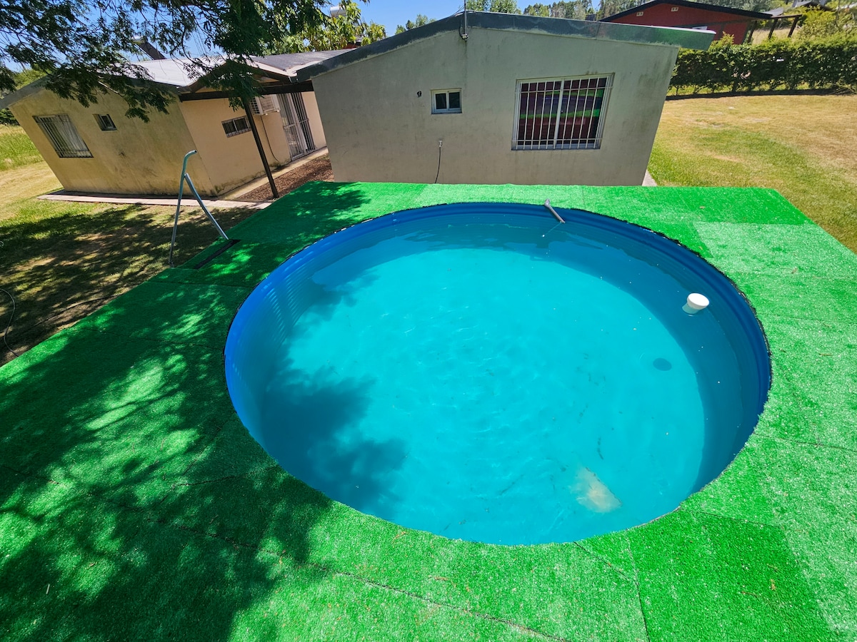 A round pool, surrounded by vibrant green grass, reflects the sunlight. A flat area is visible around the pool, contributing to an inviting outdoor space. A simple building is seen in the background, providing context to the serene environment.