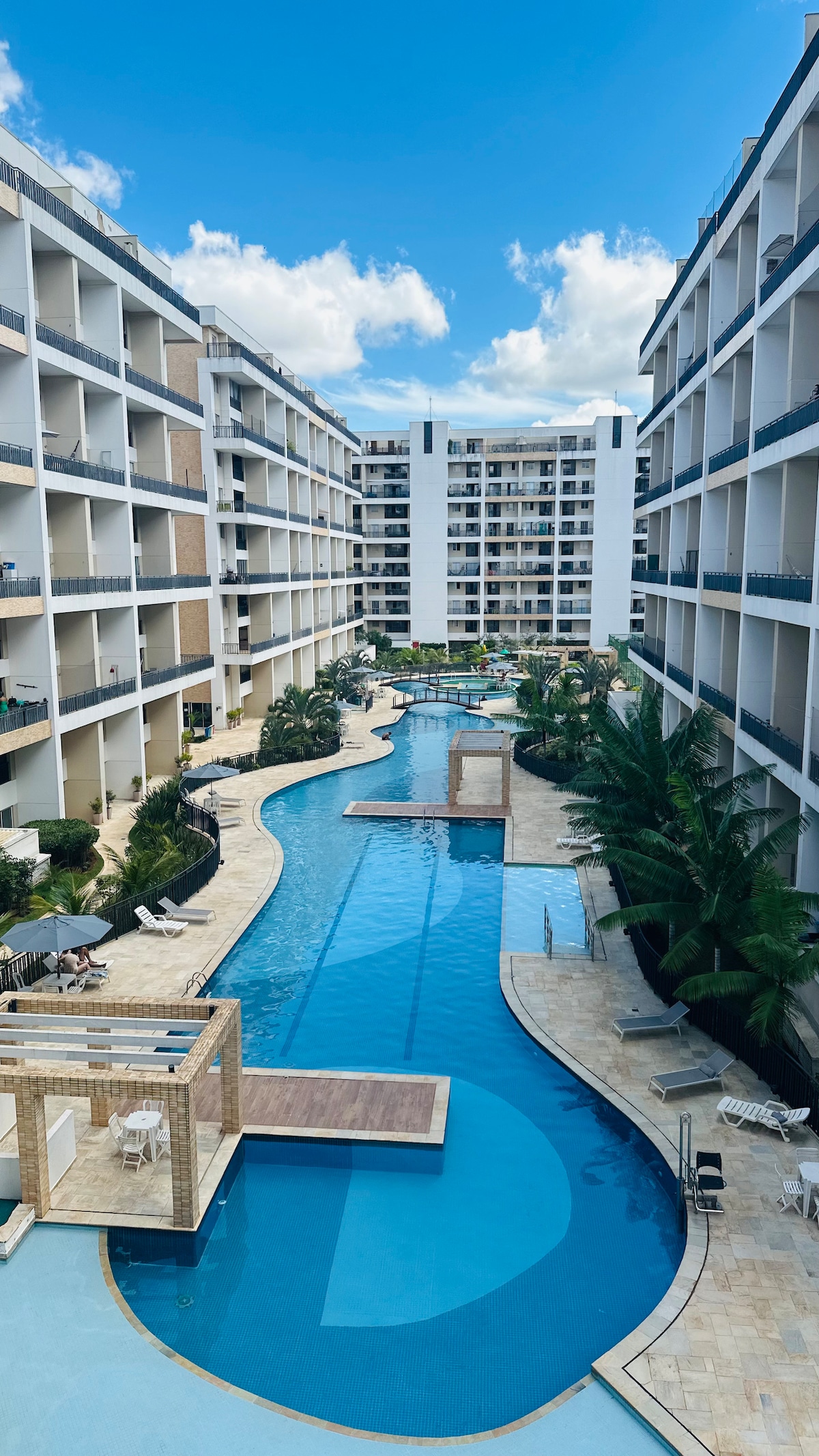 The image captures a serene pool area within a luxury condominium, featuring a winding lagoon-style pool lined with palm trees. Sun loungers are positioned along the water's edge, and the surrounding architecture is modern and inviting under a bright blue sky.