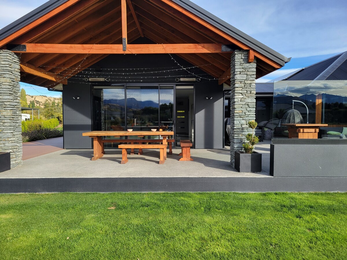 A covered outdoor area features a large wooden dining table surrounded by benches. Stone columns support a sloped roof, and glass panels offer a view of the surrounding landscape. String lights are present, creating a welcoming atmosphere for outdoor gatherings.