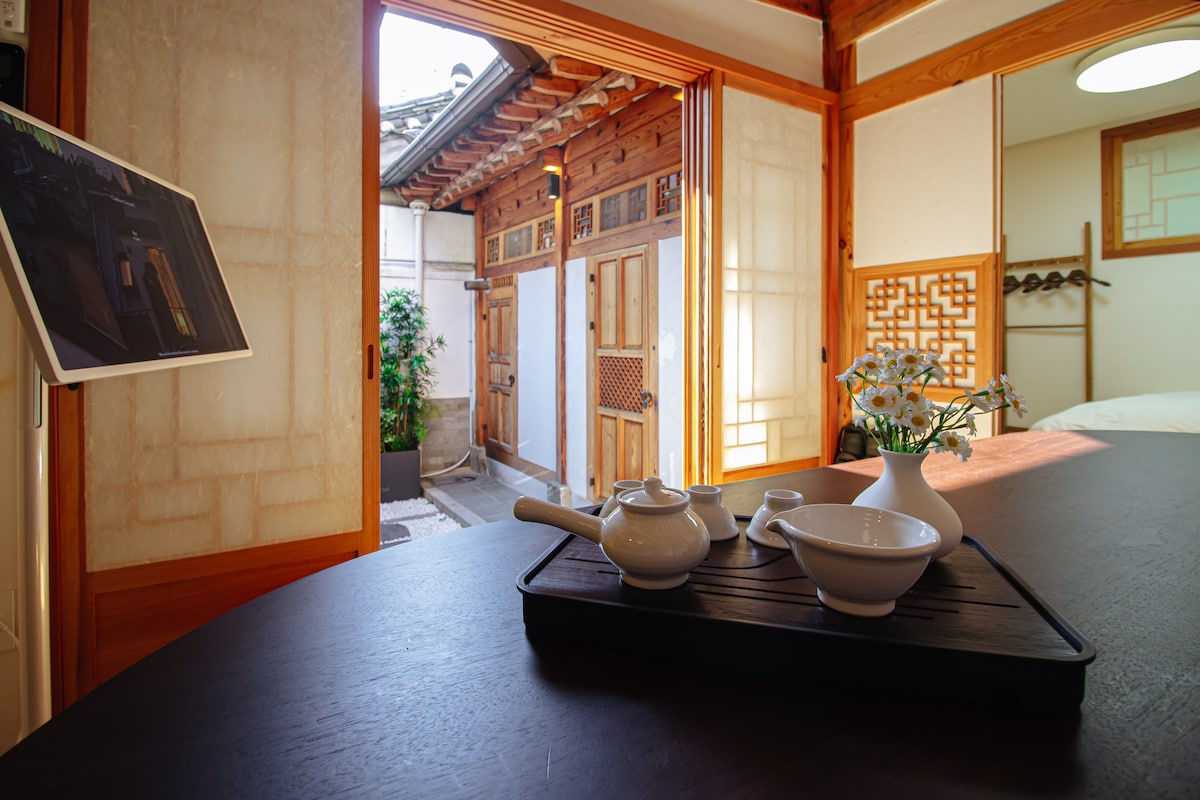 A tranquil interior space features a wooden table with a teapot and a bowl resting on a tray. Natural light is embraced through an open door, revealing a small garden area outside. The soft textures of the walls highlight the traditional architecture.
