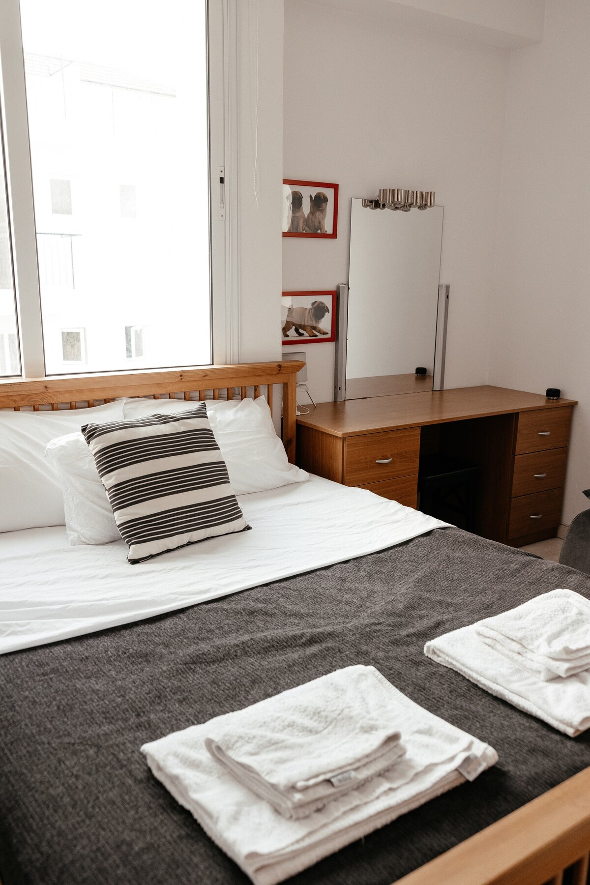 A well-appointed bedroom features a spacious bed layered with a soft grey blanket and decorative striped pillow. Two folded towels are neatly arranged at the foot of the bed. A wooden desk and mirror sit against the wall, with natural light filtering through a large window.