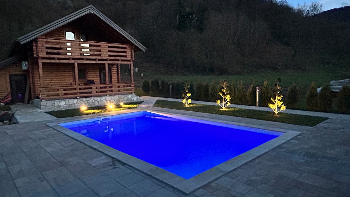 A wooden cabin is situated beside a glowing blue swimming pool, which is illuminated by subtle lighting. Lush greenery encircles the area, and a paved patio extends around the pool, enhancing the outdoor space for relaxation and gatherings.
