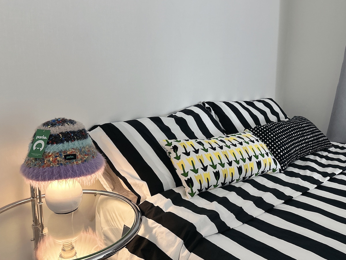 A sleeping area is presented with bold black and white striped bedding, complemented by a colorful pillow with a whimsical pattern. A stylish lamp with a fuzzy shade adds a touch of charm, positioned on a round glass side table beside the bed.