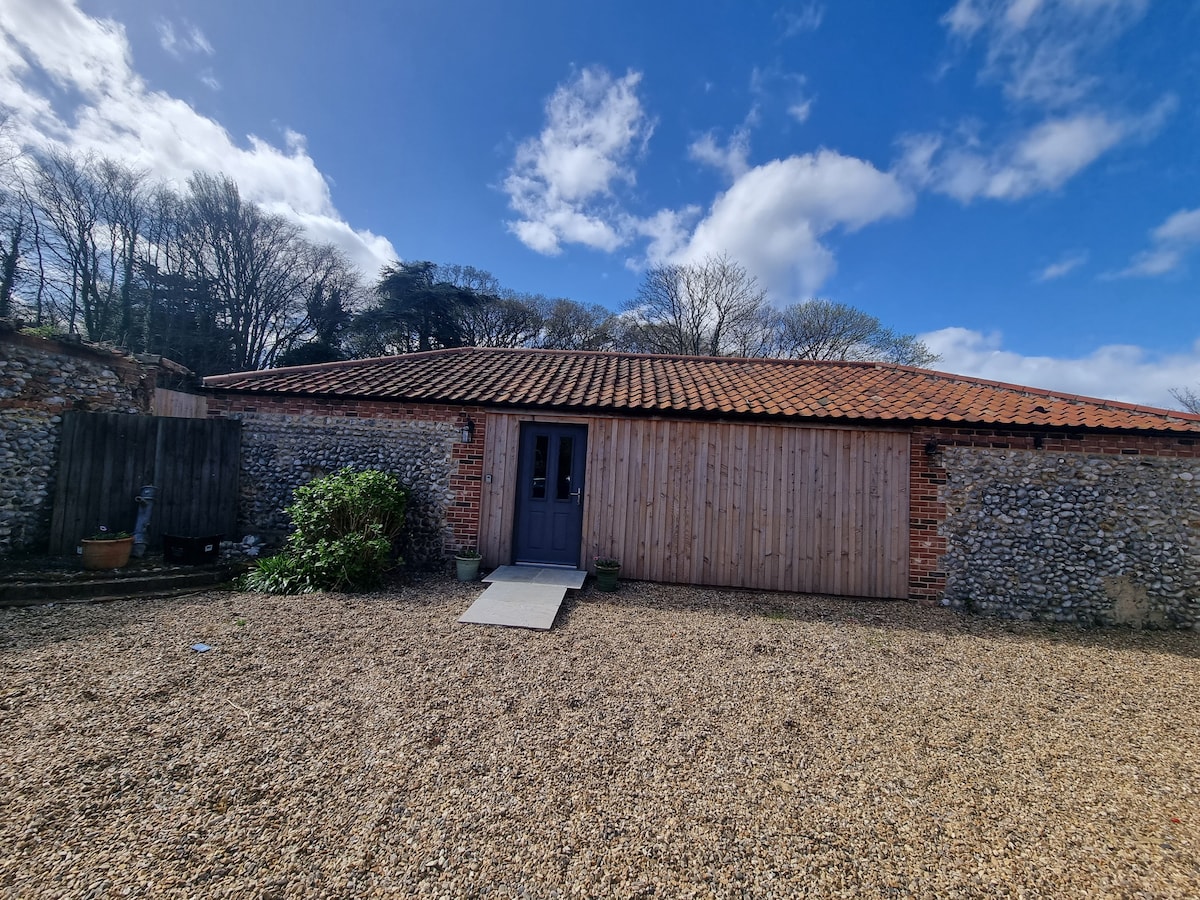 The exterior of the converted Armoury/Barn features a combination of stone and timber walls, with a red-tiled roof. Large double doors framed in dark wood open into the property. A gravel pathway leads to the entrance, surrounded by a landscaped area with trees in the background.