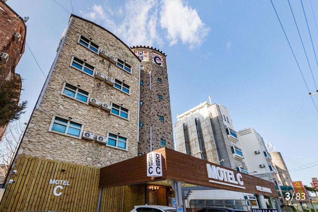 The exterior of a modern motel features a unique stone facade and multiple levels. Large windows provide natural light, while air conditioning units are visible. The entrance is marked by a prominent sign displaying the name, and the surrounding area includes additional buildings.