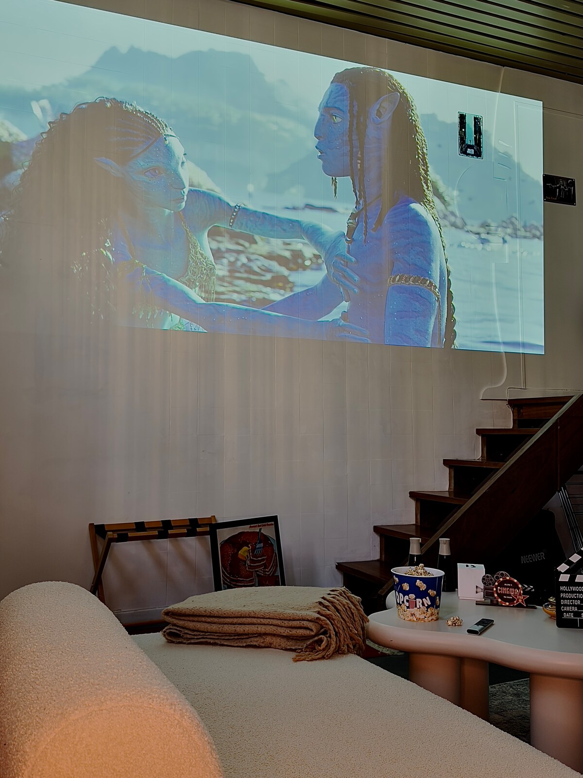 A large image projects onto a wall, displaying a scene from a film. Below the projection, a plush sofa and a cozy throw blanket provide seating. A bowl of popcorn and a small table complete the setup, creating an inviting atmosphere for a movie night.