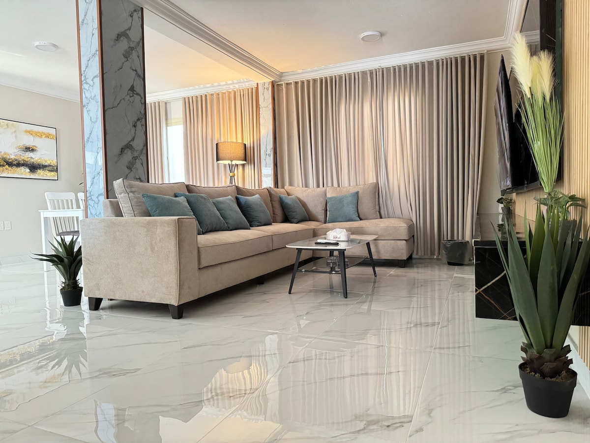 A modern living area features a spacious beige sectional sofa with decorative pillows, positioned near large windows covered by elegant sheer curtains. A stylish coffee table sits in front, complemented by tropical plants placed in decorative pots. A flat-screen TV is mounted on the wall.
