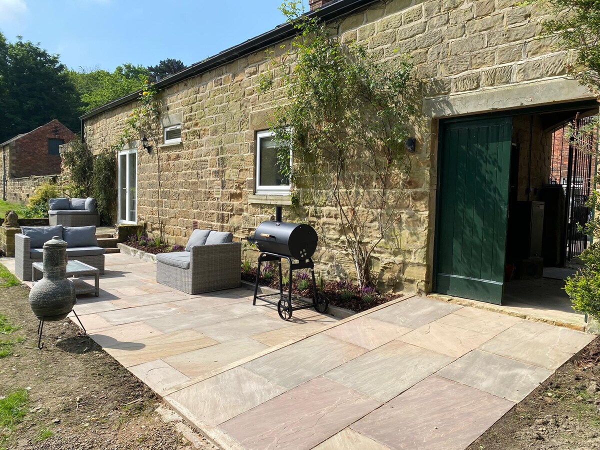 The outdoor terrace features a stone-paved area with comfortable seating and a BBQ, surrounded by greenery. Two large windows provide natural light, and the entrance leads to the interior of the cottage, enhancing connectivity between indoor and outdoor spaces.