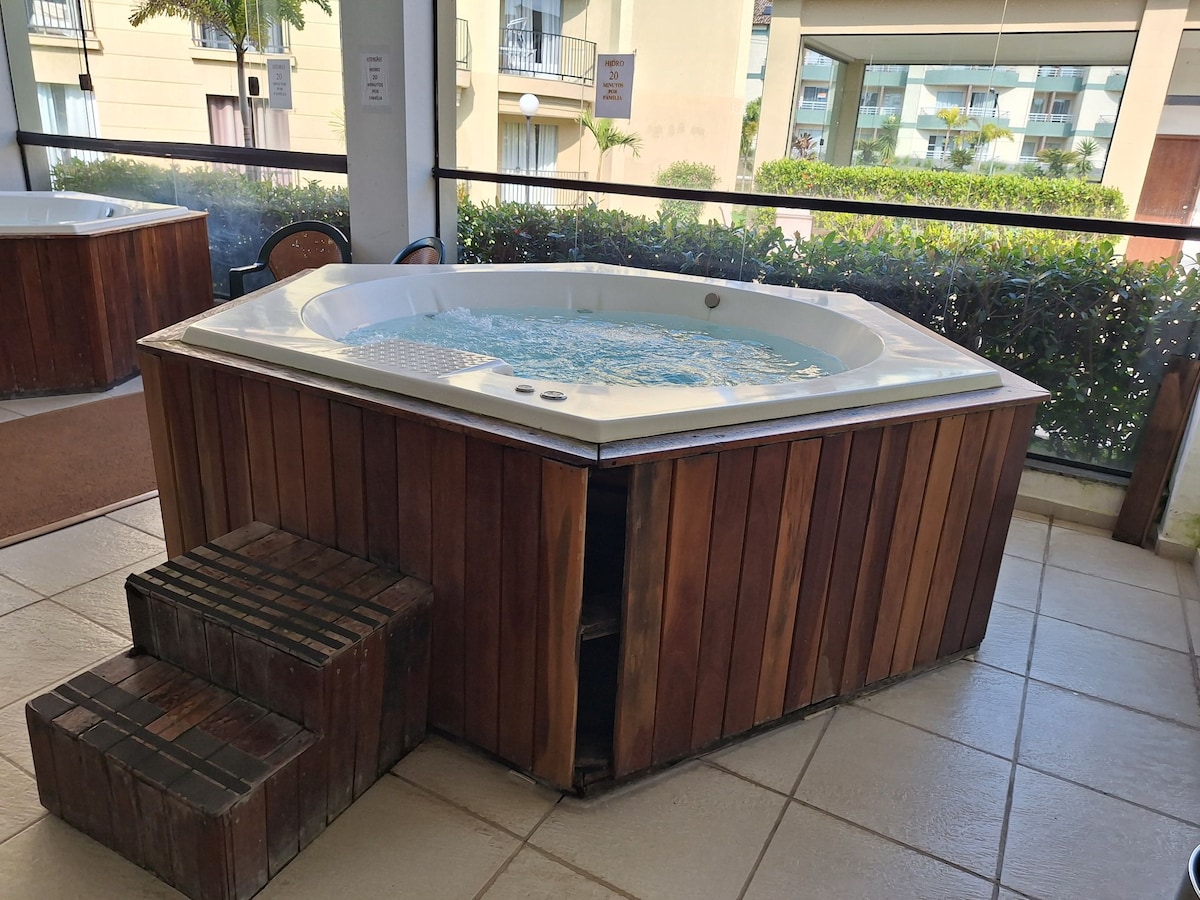 A hot tub is featured in a spacious area, surrounded by wooden panels. Steps lead up to the tub, which is filled with bubbling water. Large windows allow natural light to illuminate the space and provide views of greenery outside.
