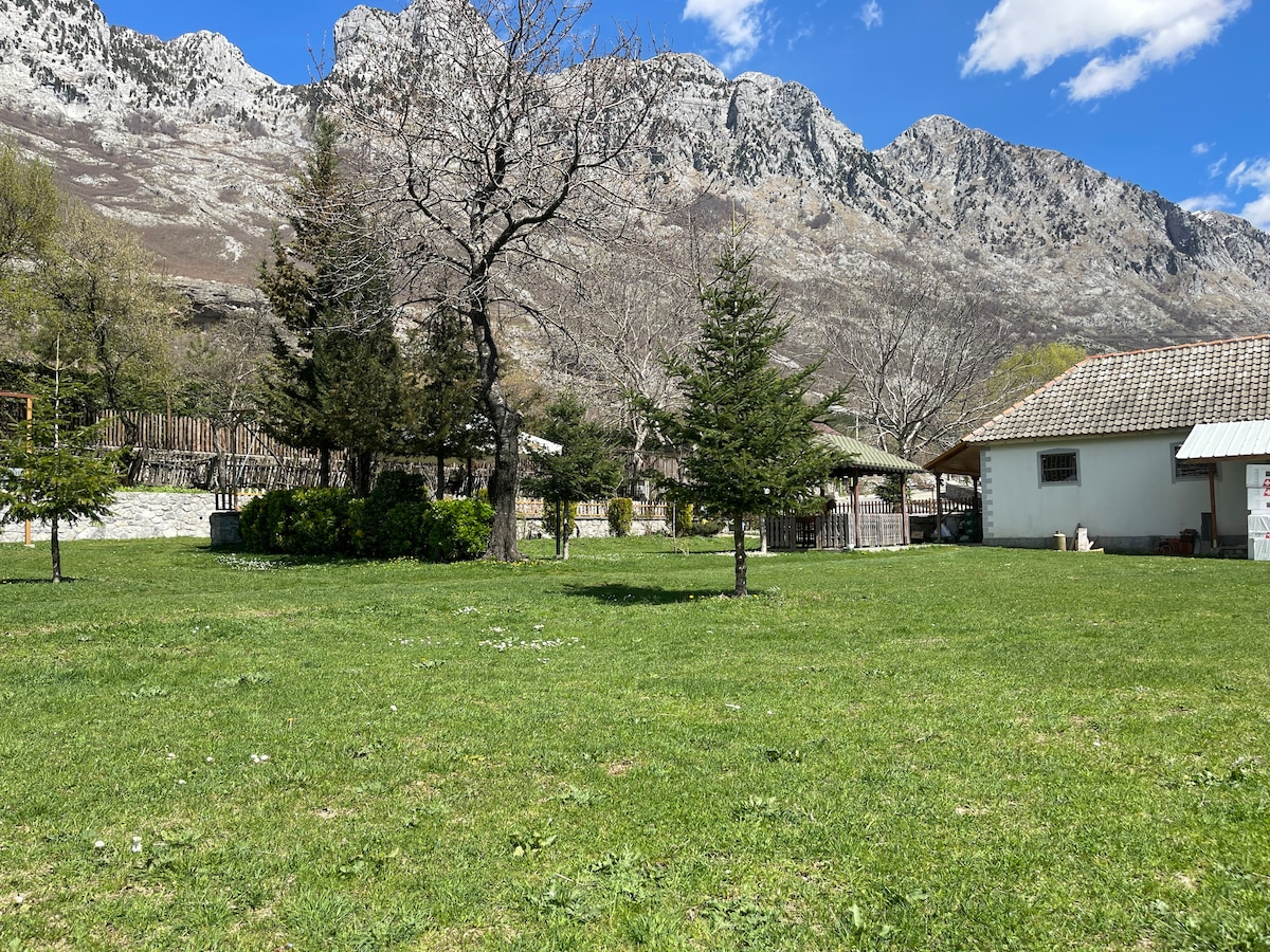 A spacious grassy area is surrounded by trees and the backdrop of towering mountains. Two buildings are visible, showcasing a traditional architectural style, while open space encourages outdoor activities. The clear sky adds brightness to the serene landscape.