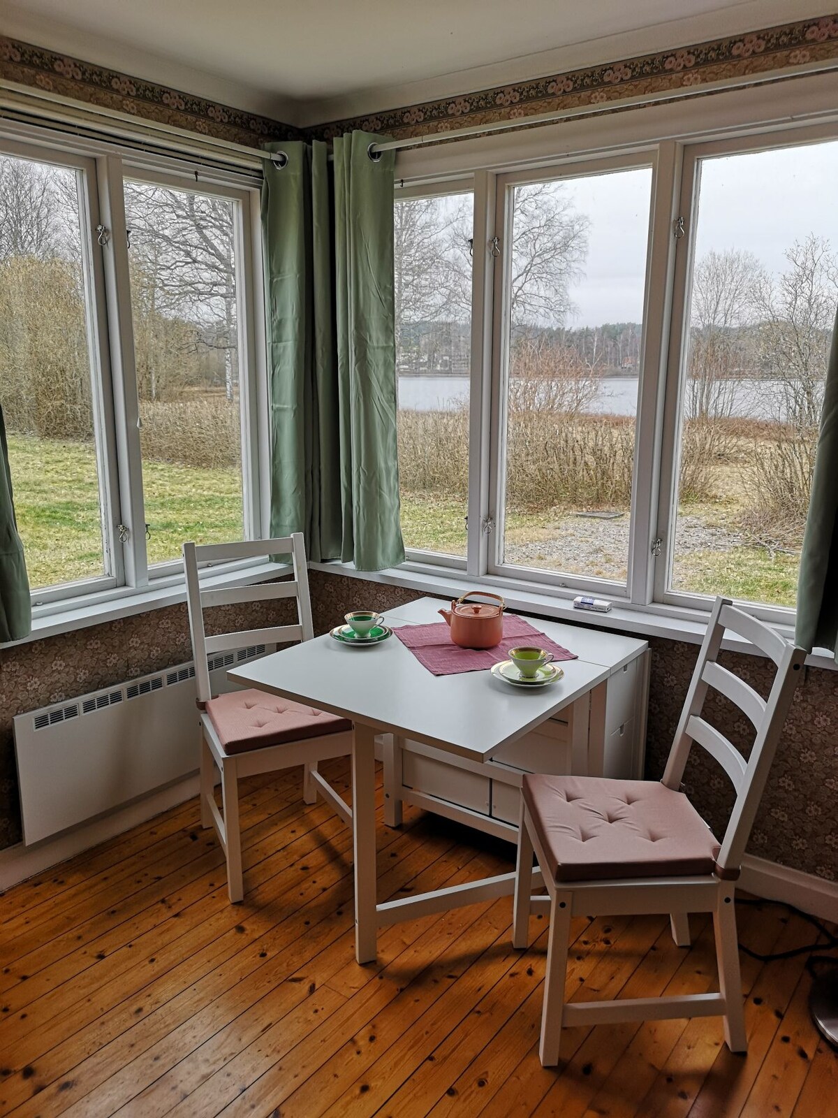 A small dining table with two chairs is situated beside large windows, offering a view of the lake and surrounding greenery. Light green curtains frame the windows, enhancing the natural light in the space. A simple place setting is arranged on the table.