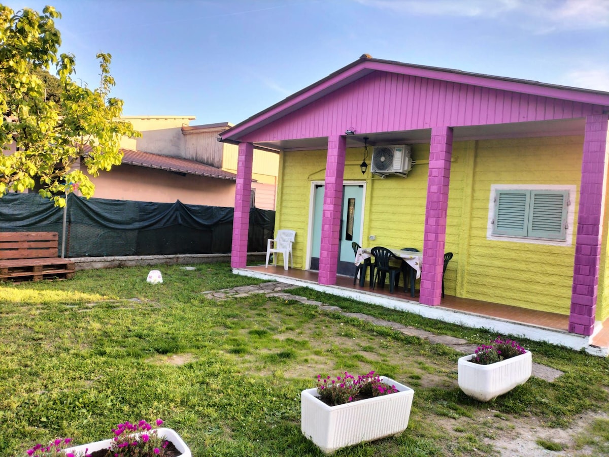 A family-friendly space is showcased, featuring a colorful exterior with yellow and pink walls. A covered porch includes black chairs and a table. Lush green grass surrounds the building, highlighted by white flower planters. A shaded area is visible in the background.