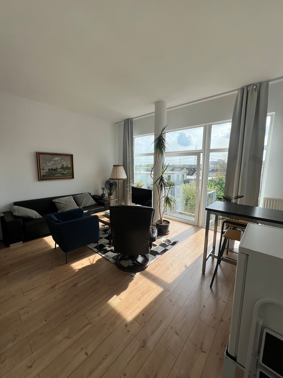 A bright living area features large windows allowing natural light to illuminate the space. Comfortable seating, including a dark sofa and two armchairs, surrounds a central table. A tall plant adds a touch of greenery, while a lamp provides additional lighting.