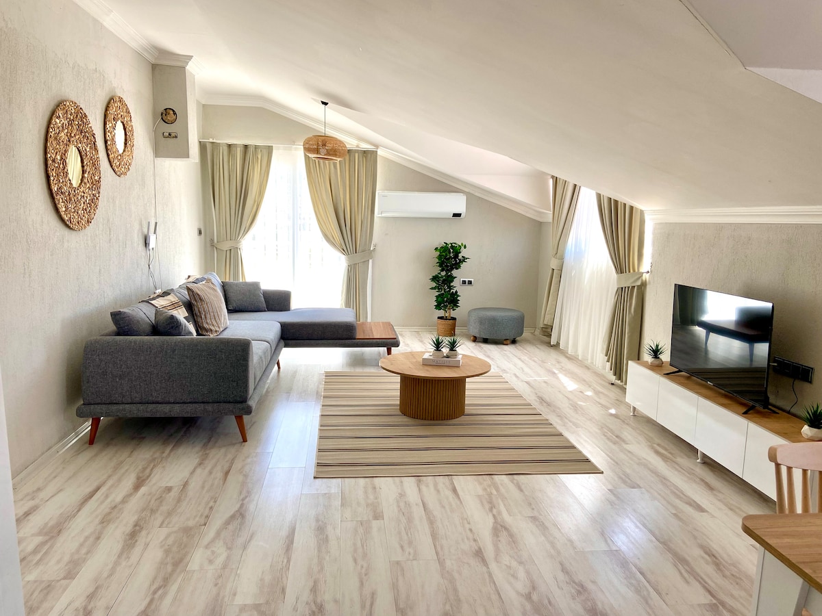 A spacious living area features a comfortable grey sofa accompanied by a round coffee table. Soft natural light filters through large windows with flowing curtains. A flat-screen TV is positioned on a modern white cabinet, while decorative plants add a touch of greenery.