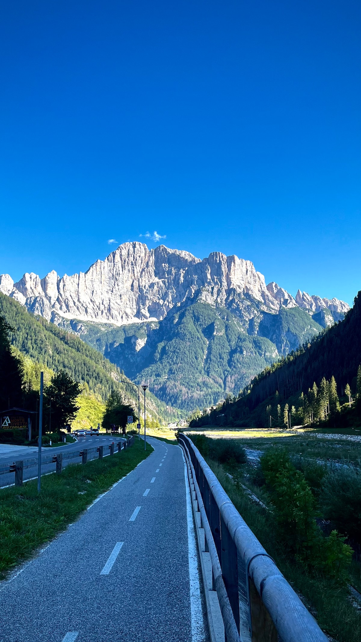 A scenic pathway is flanked by greenery, stretching alongside a river and leading towards majestic mountains. The clear blue sky enhances the vibrant colors of the landscape, with rocky peaks visible in the background rising dramatically against the horizon.