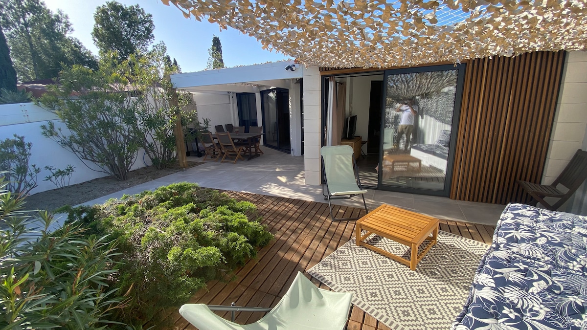A spacious outdoor area features wooden decking with a small table and chairs, surrounded by greenery. A shaded seating space is provided by a fabric canopy, offering a cozy setting for relaxation. Large glass doors open into the interior, enhancing the connection between indoor and outdoor spaces.