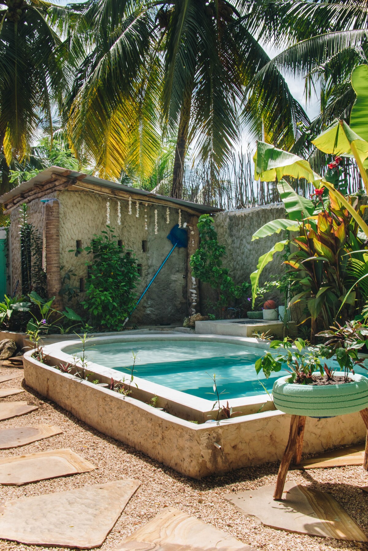 A private pool is framed by lush tropical plants and palm trees, providing a relaxing outdoor space. The area features stone pathways leading to the pool, enhancing the tranquil atmosphere. A bamboo structure and various green plants contribute to the natural aesthetic surrounding the pool.