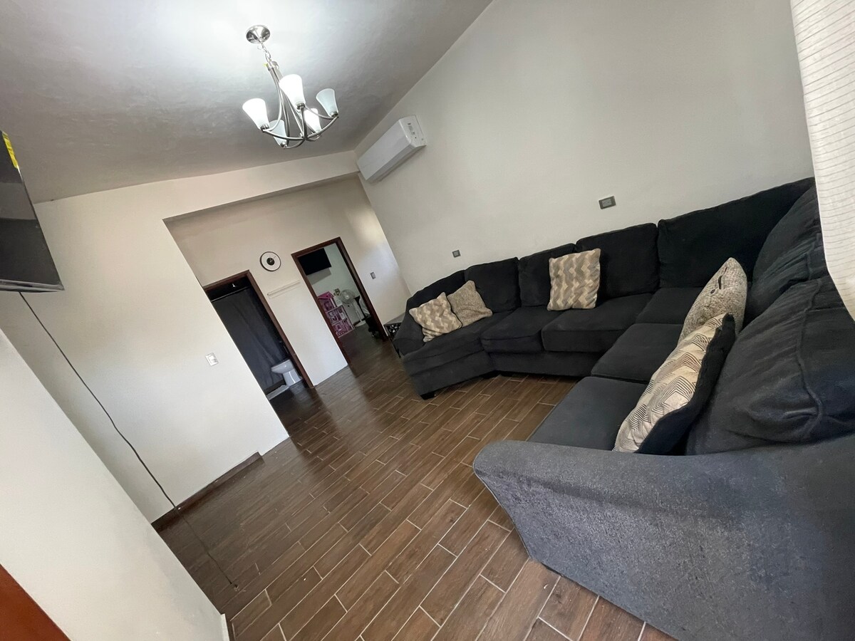 A spacious living area is furnished with a large sectional sofa adorned with textured pillows. A flat-screen television is mounted on the wall, and a ceiling fan provides airflow. Light-colored walls enhance the feeling of openness, with natural light filtering in through a nearby doorway.