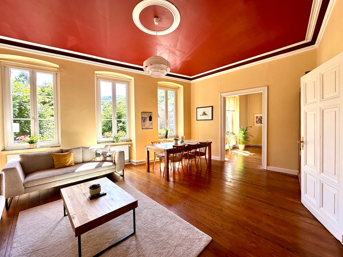 A spacious living area is highlighted by a deep red ceiling and warm beige walls. Large windows allow natural light to illuminate the room, featuring a comfortable sofa and a dining table surrounded by chairs. A coffee table sits on a soft area rug, adding to the inviting atmosphere.
