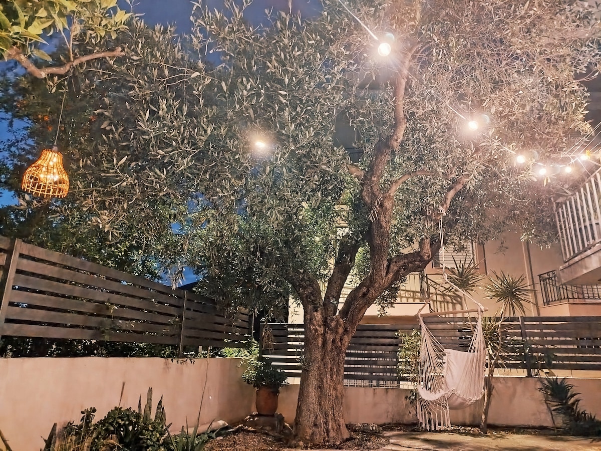 An olive tree stands prominently in a serene outdoor space, surrounded by softly glowing string lights. A comfortable hammock is suspended nearby, enhancing the relaxed atmosphere. Wooden slats create a boundary while various plants add greenery to the inviting setting.