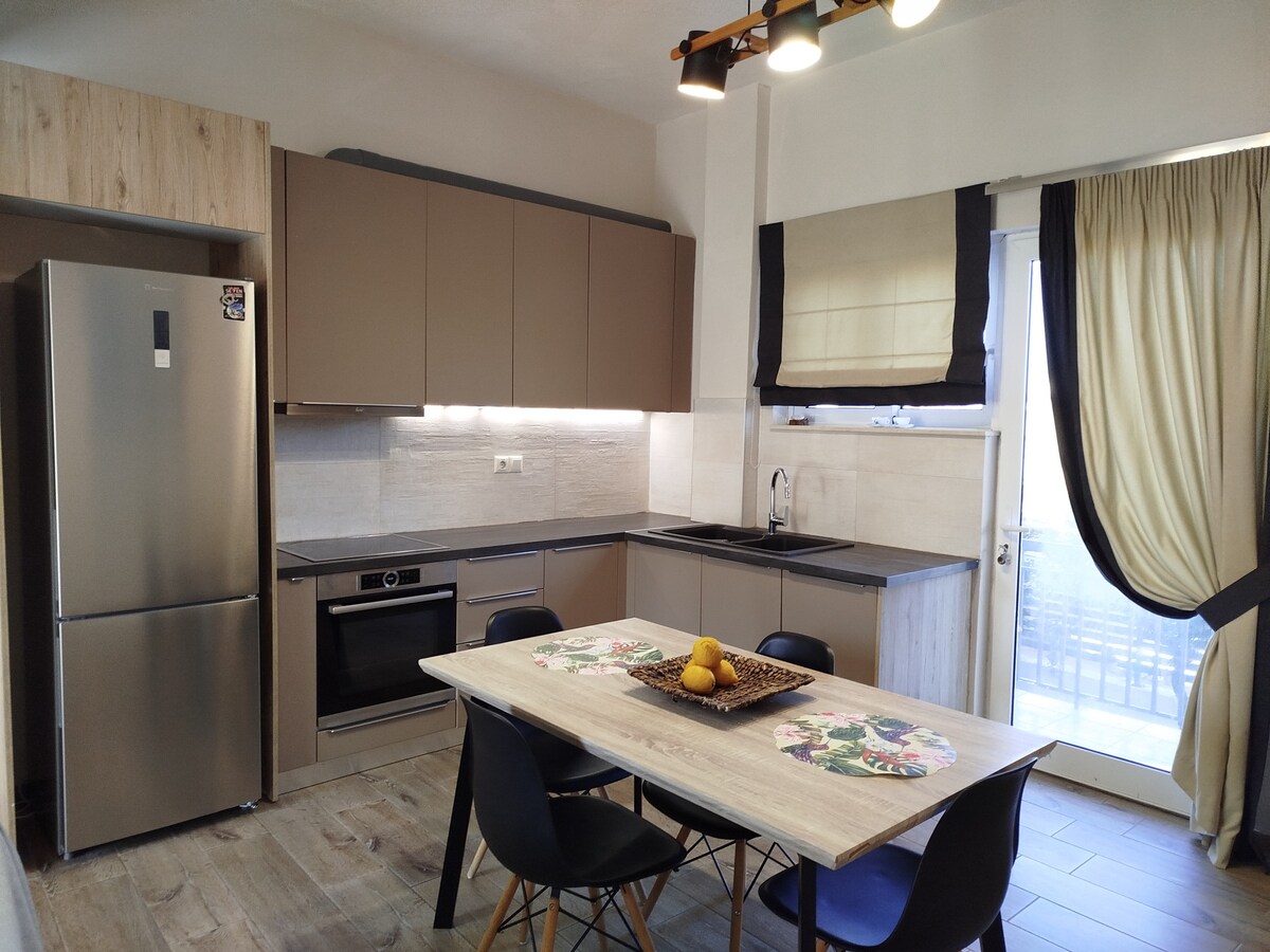 A modern kitchen is presented with sleek cabinetry and a spacious countertop. Equipped with a stainless steel refrigerator, an oven, and a sink, the kitchen facilitates cooking and dining. A wooden table with black chairs is set, featuring a decorative bowl of fruit.