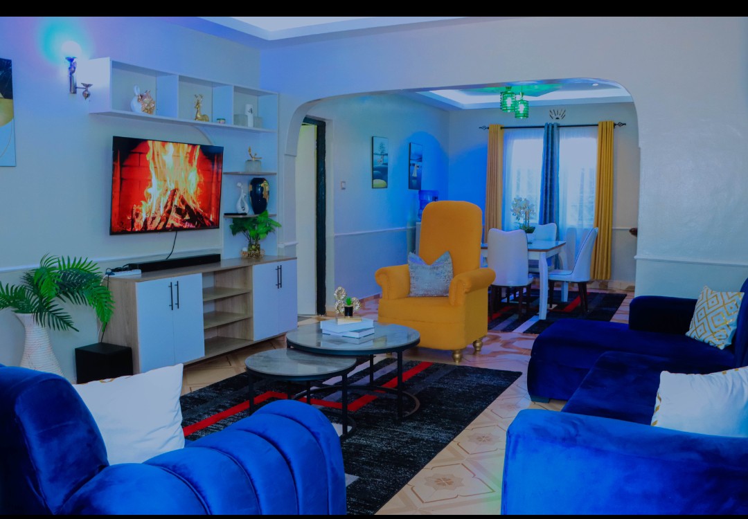 A modern living area is presented with a large television displaying a fireplace scene. Comfortable blue sofas are arranged around a coffee table, complemented by a decorative rug. Bright window coverings allow natural light to fill the space, enhancing the overall ambiance.