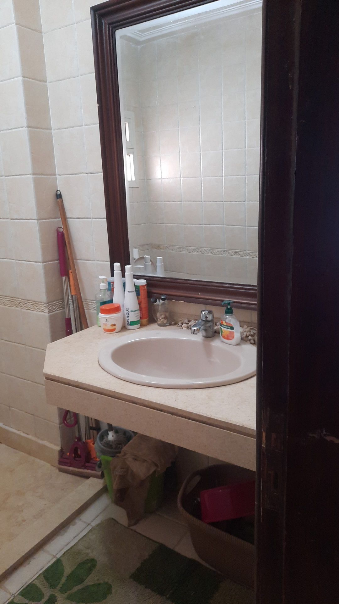 A compact bathroom featuring a light-colored sink with a mirror above it. Various toiletries are neatly arranged near the faucet. The tiled walls are complemented by a subtle decorative border. Items are visible on the counter, and a storage basket is placed on the floor.