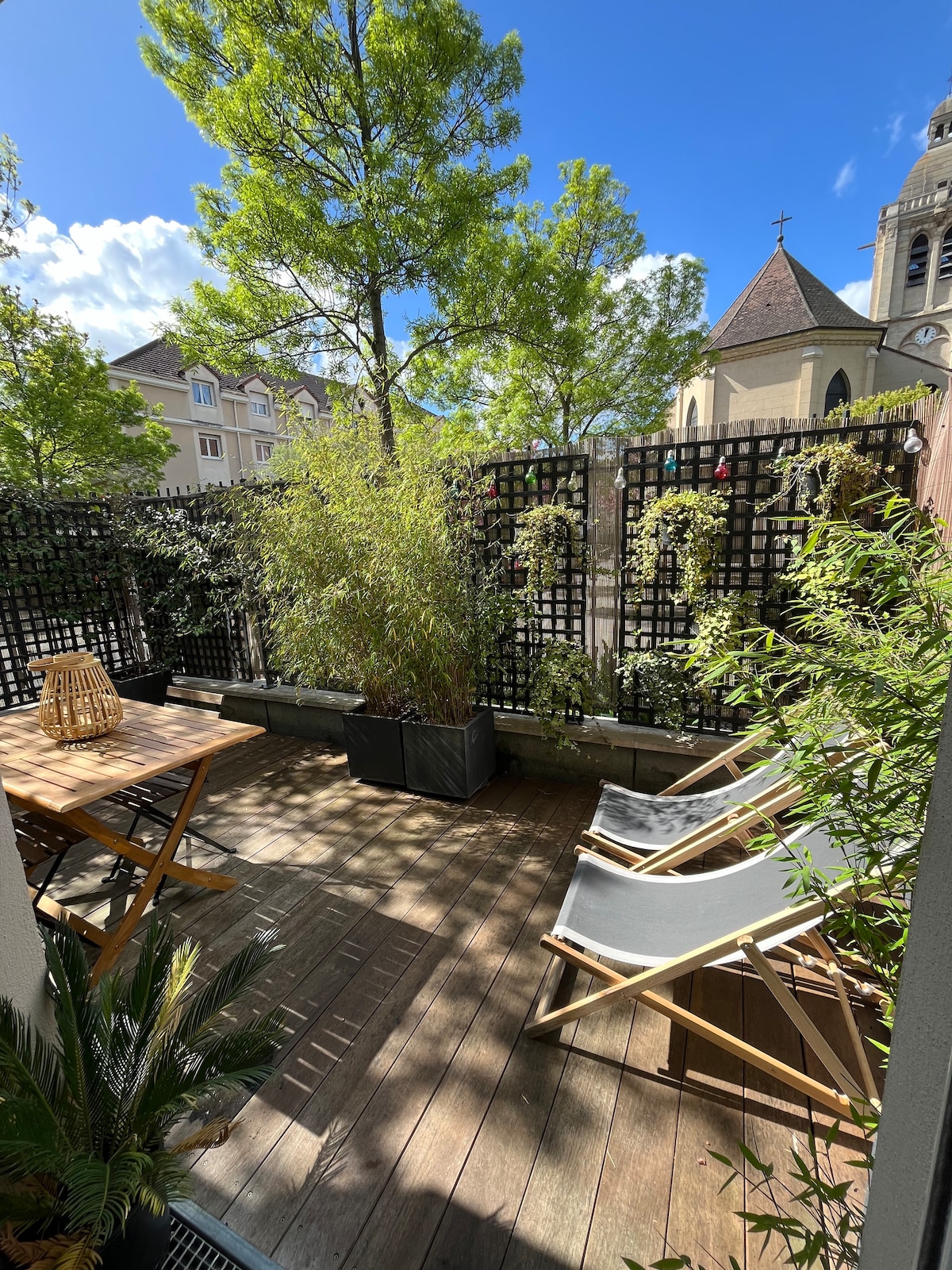 A verdant terrace is showcased, featuring wooden flooring and surrounded by lush greenery. A small wooden table and chairs provide seating, while two lounge chairs invite relaxation. The backdrop includes decorative trellises and glimpses of an adjacent building and church steeple.