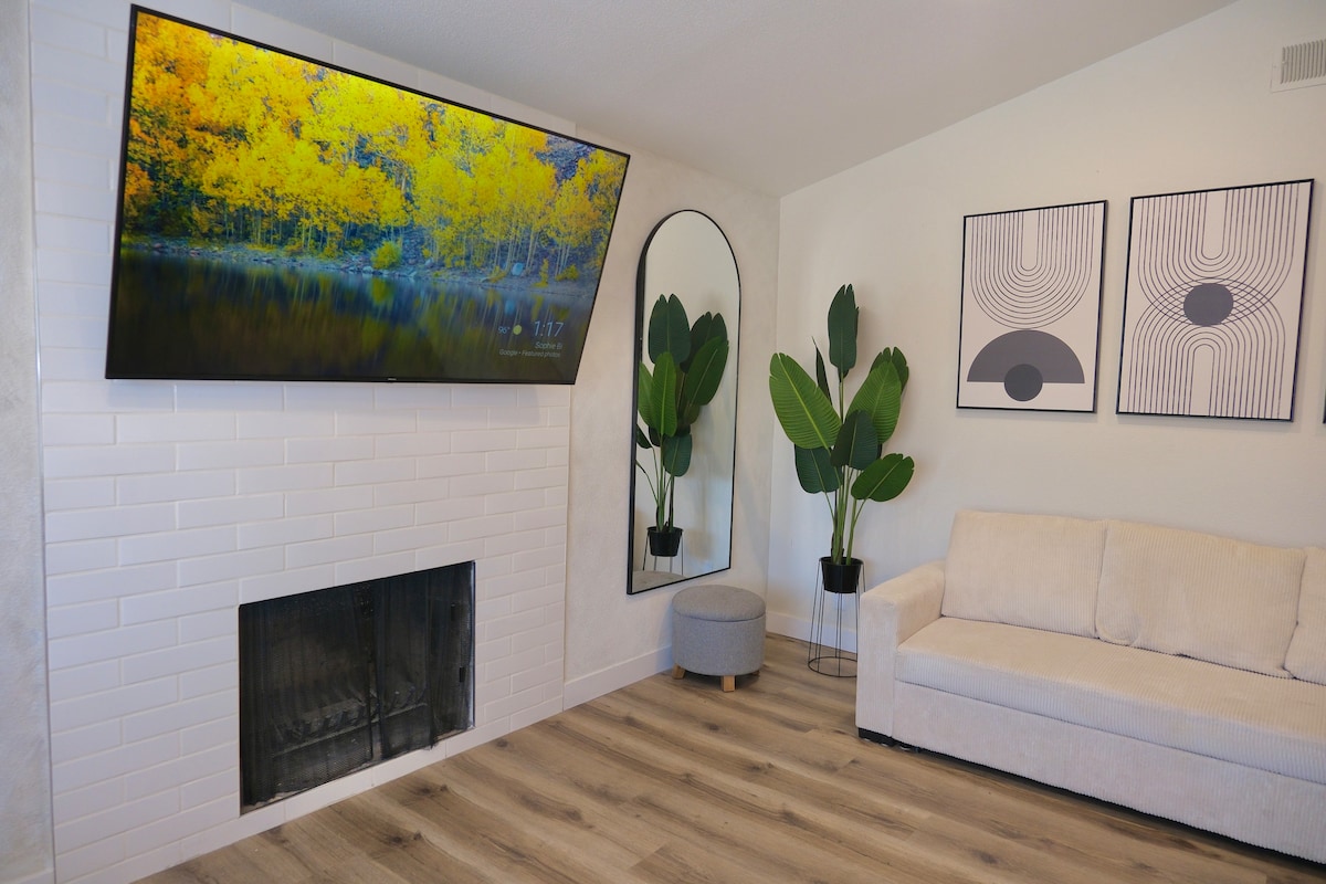 A modern living area is highlighted by a large flat-screen TV mounted above a white tiled fireplace. Two framed artworks are displayed on the wall. A round ottoman and a light-colored sofa provide comfortable seating, while potted plants add a touch of greenery.