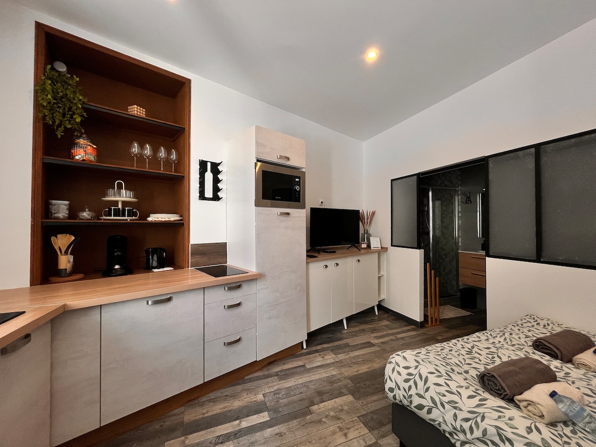 A modern studio kitchen area is equipped with sleek cabinetry and essential appliances, including a microwave and coffee maker. Adjacent to the kitchen, a cozy sleeping space features a patterned blanket over the bed, with natural light illuminating the room through glass partitions.