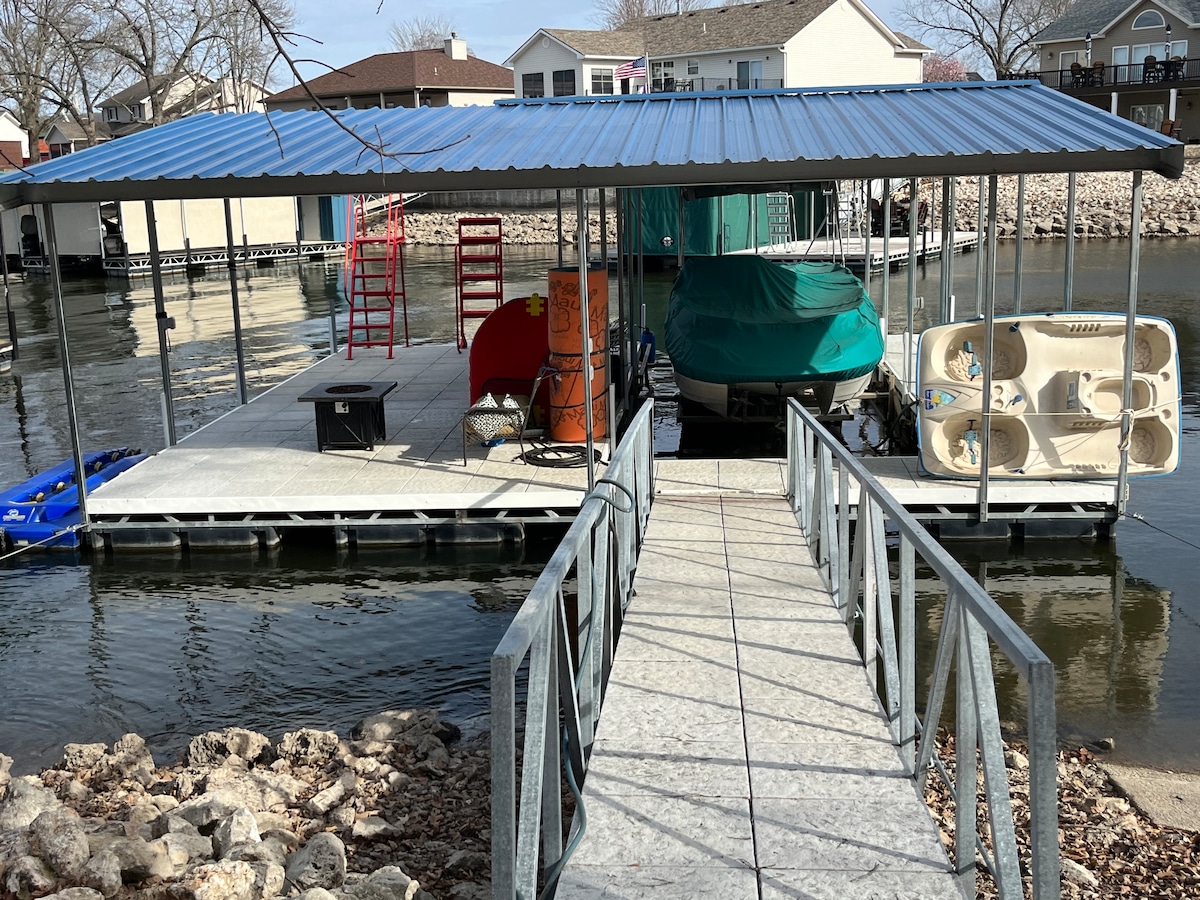 A covered dock is seen extending over calm waters, featuring various watercraft including a green boat and a paddle boat. A wide walkway leads to the dock, while equipment like chairs and a barrel is arranged nearby.